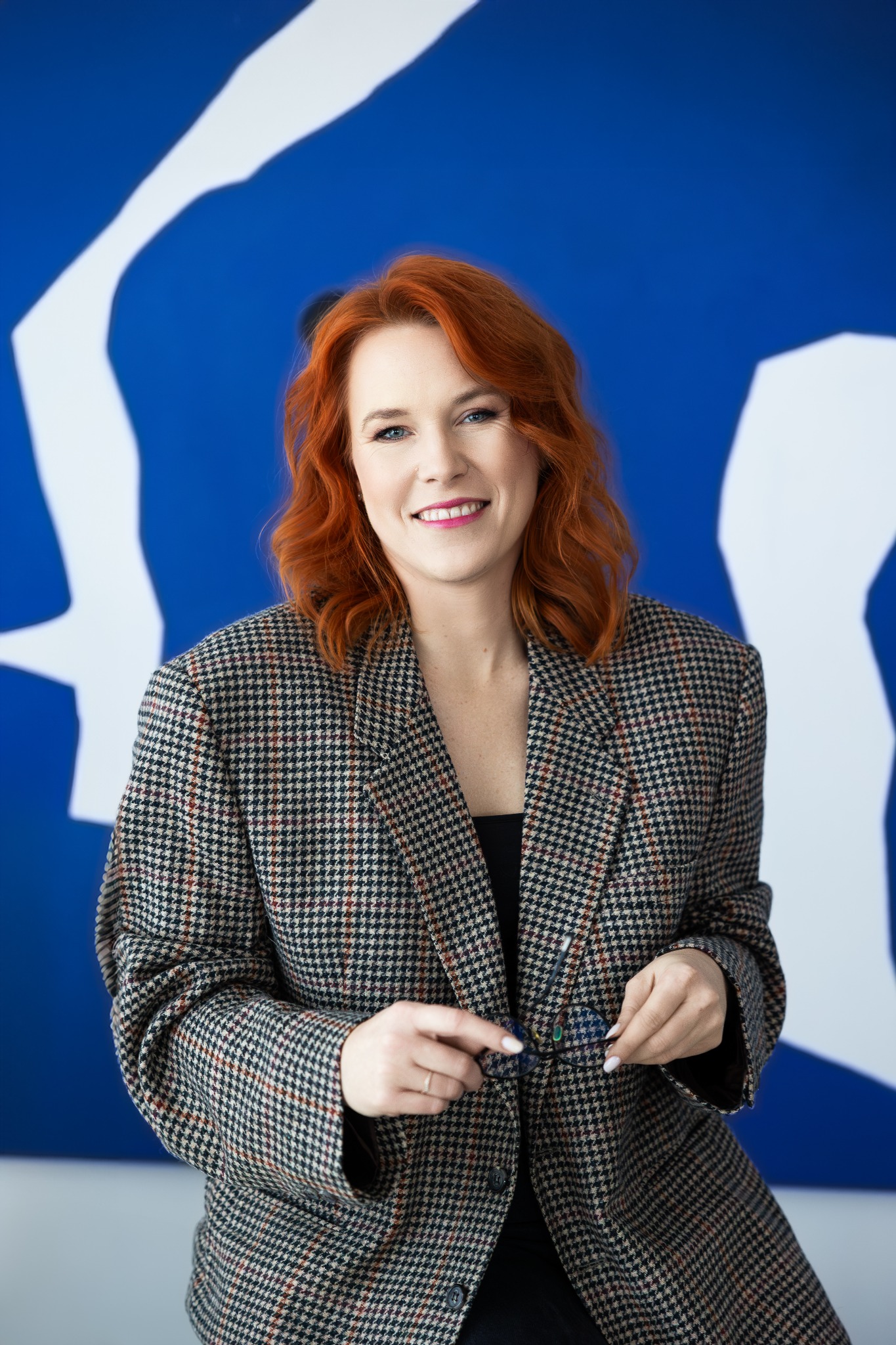 Uśmiechnięta kobieta z rudymi włosami w marynarce w kratę, trzymająca okulary na tle niebieskiej ściany z białym abstrakcyjnym wzorem. Portret biznesowy.