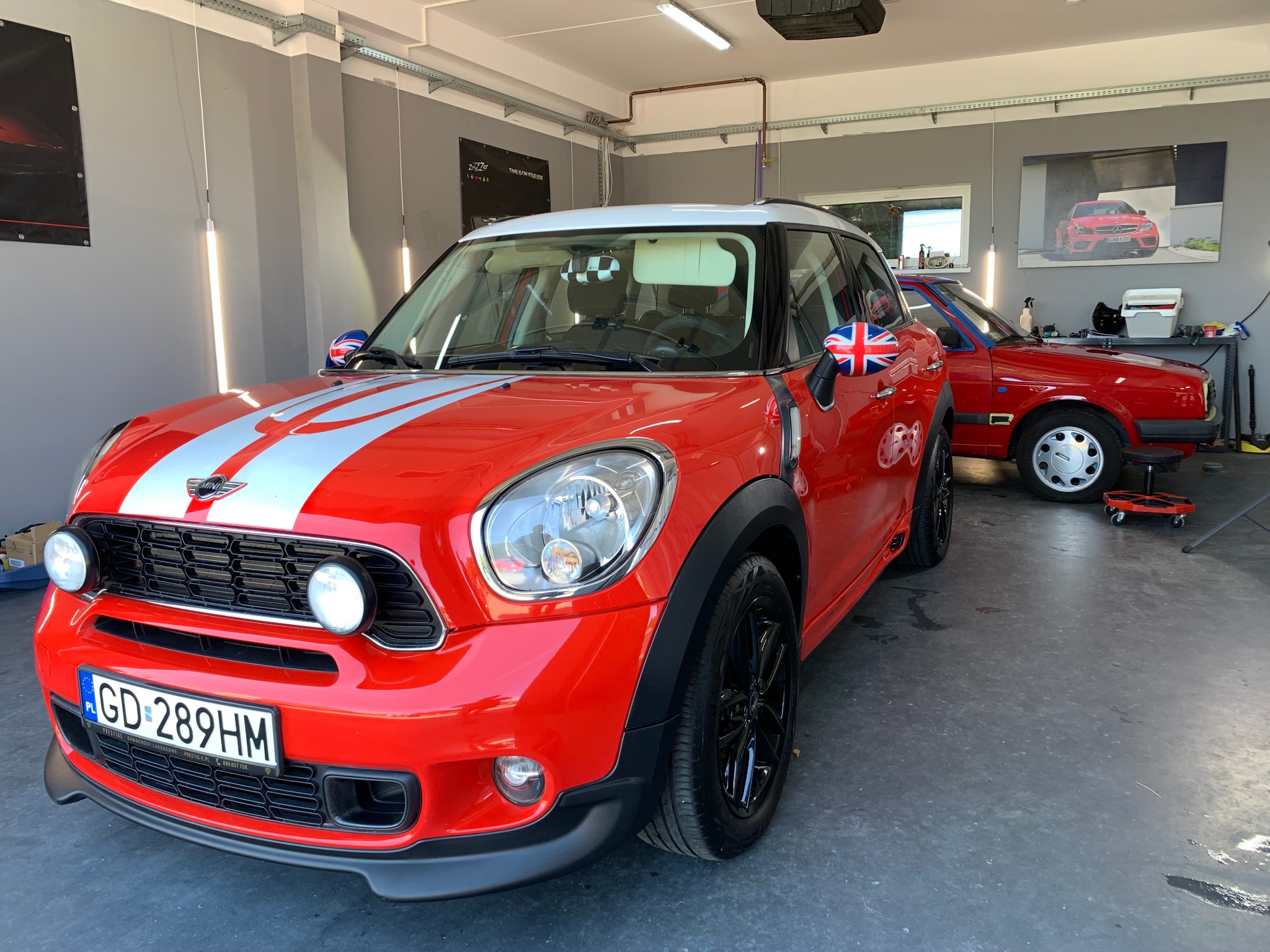 Czerwony Mini Cooper po detailingu w myjni, z flagą Wielkiej Brytanii na lusterkach, widoczny fragment drugiego auta i plakat z autem w tle.