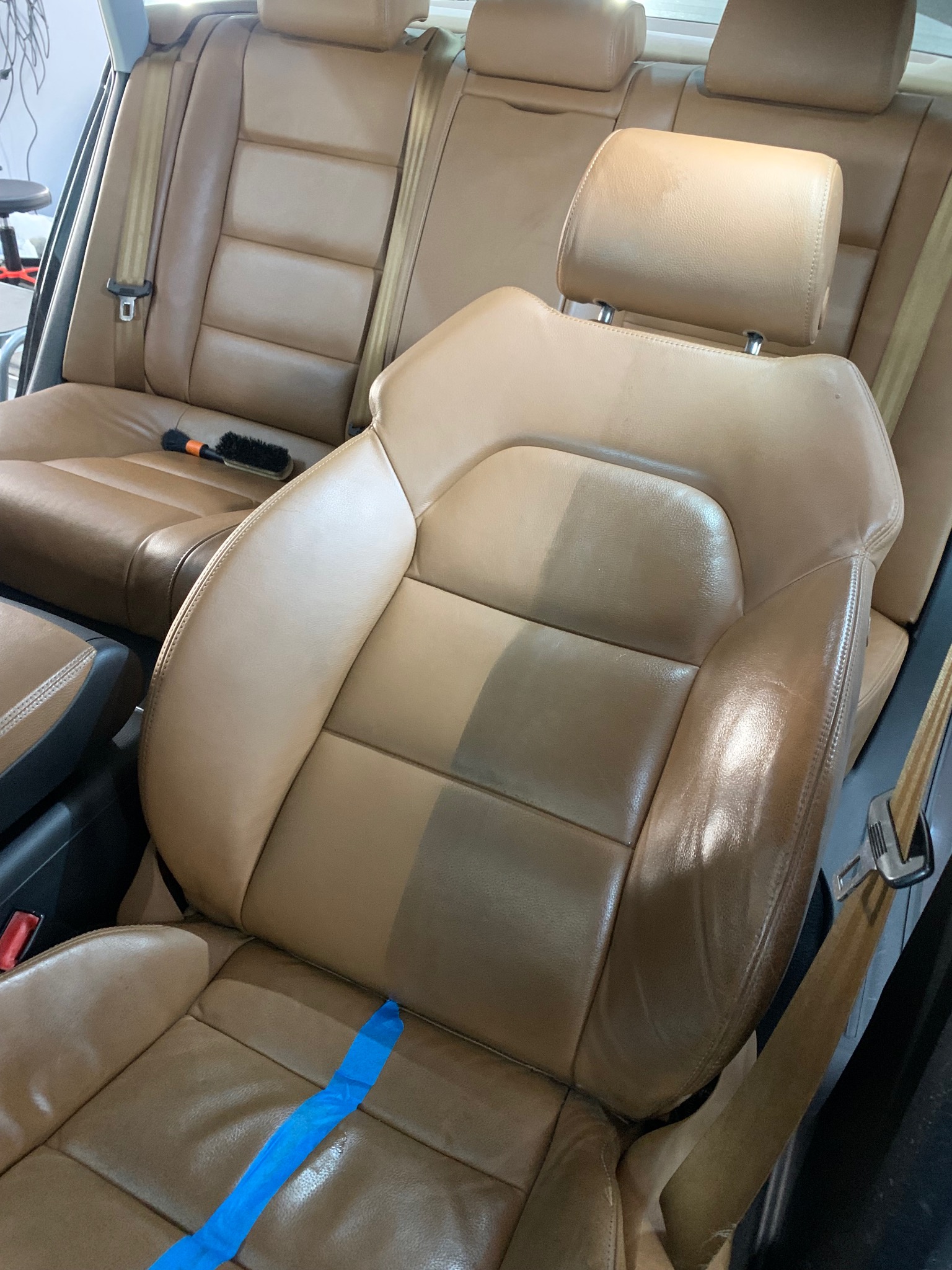 Brązowa skórzana tapicerka samochodowa, fotel kierowcy w trakcie czyszczenia: połowa wyczyszczona, druga - brudna. Widoczna taśma malarska i szczotka do czyszczenia.