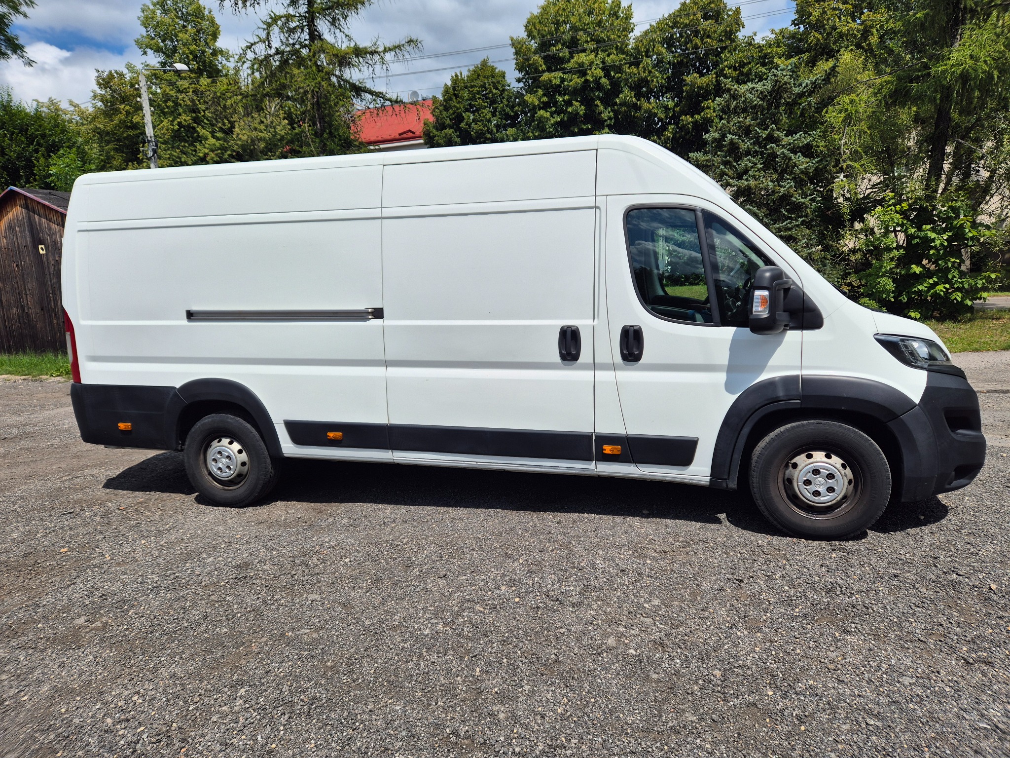 Biały bus dostawczy na żwirowym podłożu, widok z boku, zieleń w tle. Pojazd z przesuwnymi drzwiami.