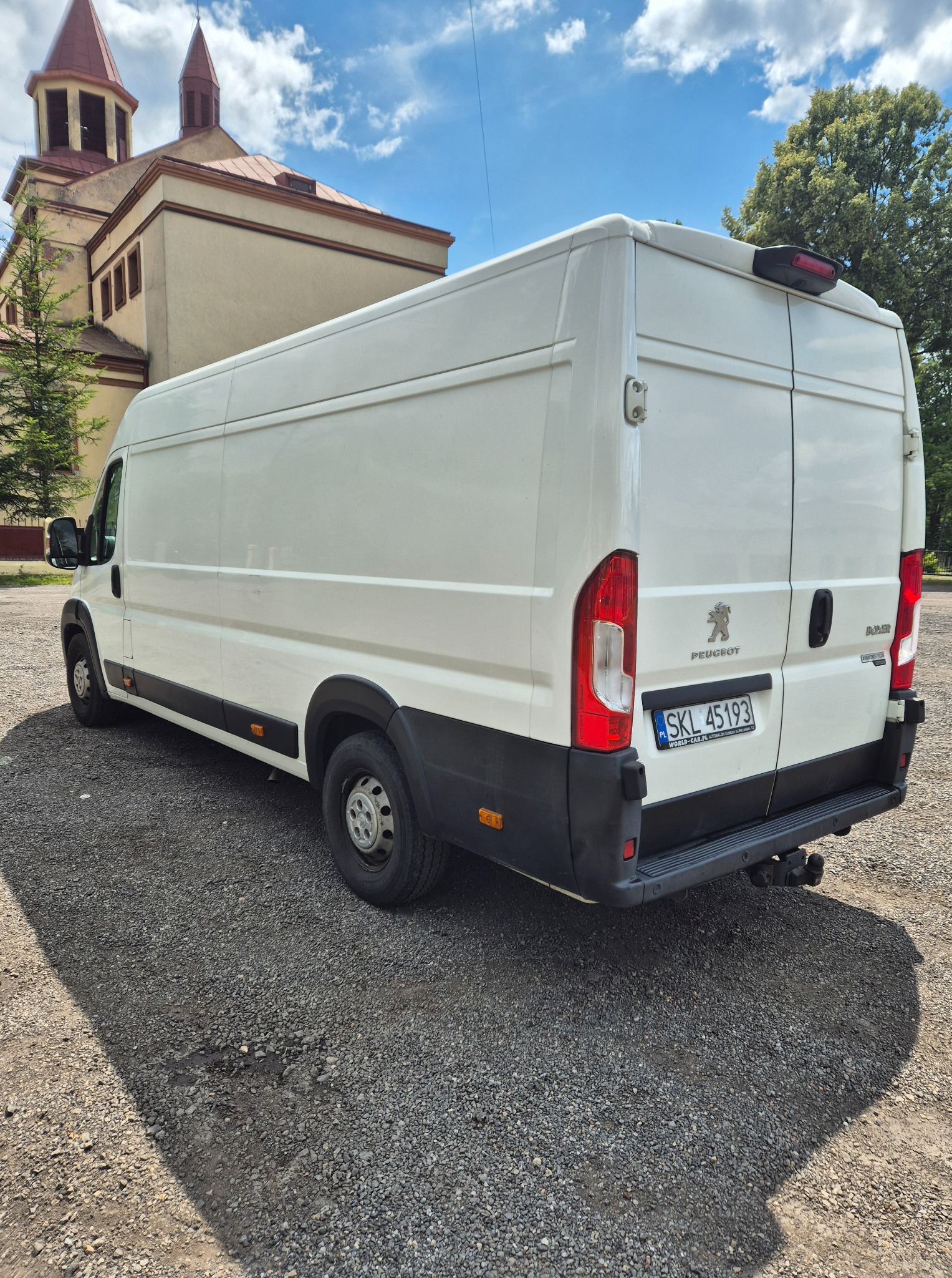 Biały bus Peugeot Boxer zaparkowany na żwirowym placu, z tyłu widoczny budynek kościoła z czerwonymi wieżami. Słoneczny dzień, widoczne cienie.