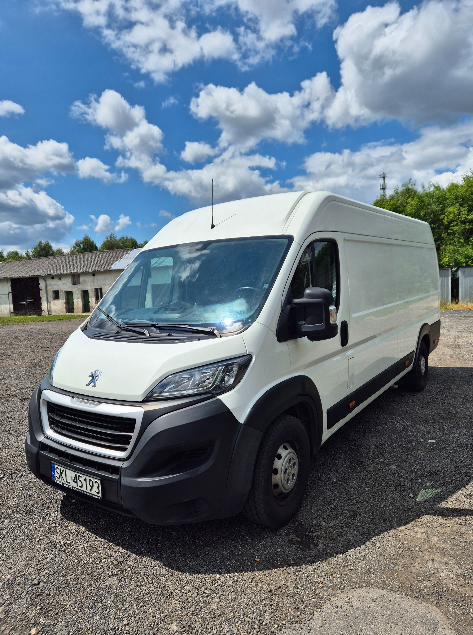 Biały bus Peugeot z rejestracją SKL 45193 na żwirowym podłożu, w tle budynek gospodarczy i błękitne niebo z obłokami. Auto z perspektywy lekko od dołu.