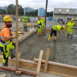 Pracownicy w kaskach wyrównują świeżą wylewkę na zbrojeniu. W tle budynki mieszkalne i plac budowy. Jeden z pracowników patrzy w obiektyw.