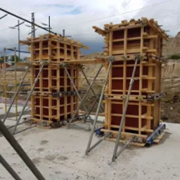 Szare podpory podtrzymują drewniany szalunek kolumn na budowie. W tle widoczne są wykopane warstwy ziemi i konstrukcja zbrojeniowa.