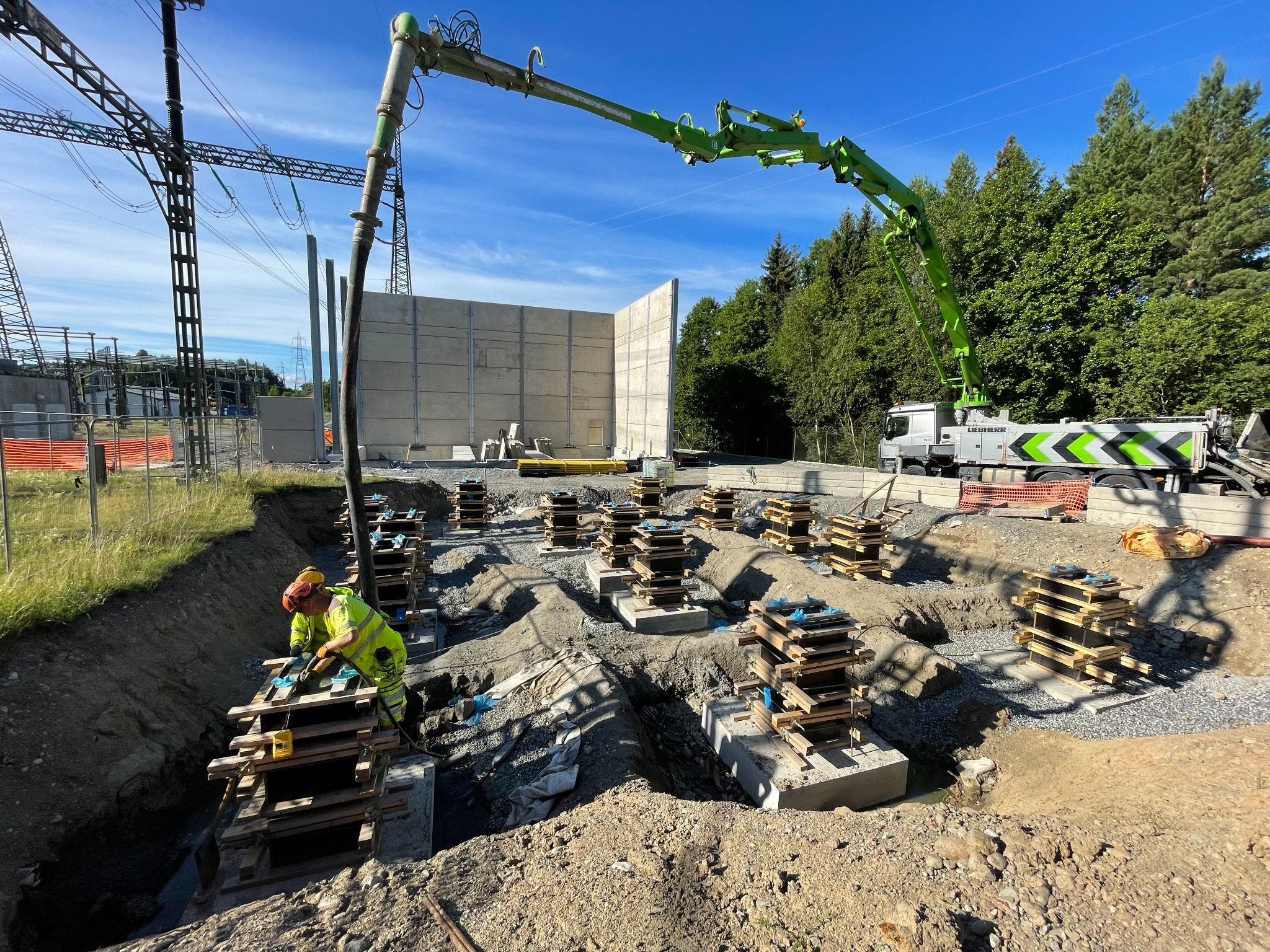 Budowa fundamentów: pracownik w żółtej kamizelce montuje szalunki, widoczna pompa do betonu i betonowe ściany w tle, teren budowy z elementami infrastruktury energetycznej.