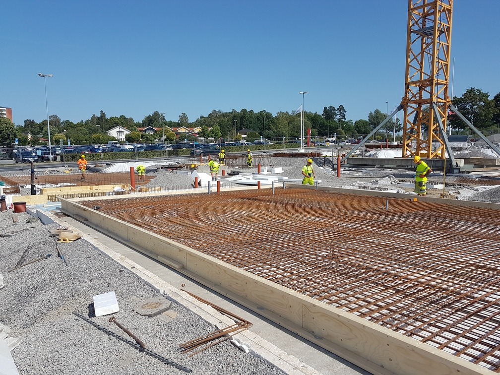 Zbrojenie fundamentu na placu budowy z widocznym żurawiem i pracownikami w odblaskowych ubraniach. Gotowa siatka zbrojeniowa w szalunku, przygotowana do zalania betonem.