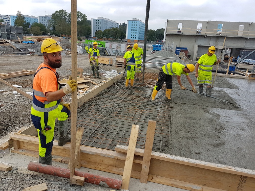 Pracownicy w kaskach wyrównują świeżą wylewkę na zbrojeniu. W tle budynki mieszkalne i plac budowy. Jeden z pracowników patrzy w obiektyw.