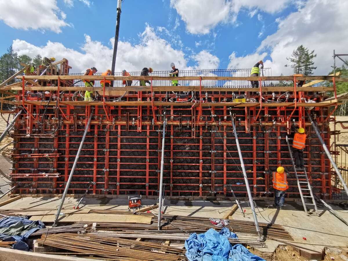 Ekipa budowlana montuje szalunek pod konstrukcję żelbetową. Widoczne elementy szalunku, zbrojenia i pracownicy w odblaskowych kamizelkach.