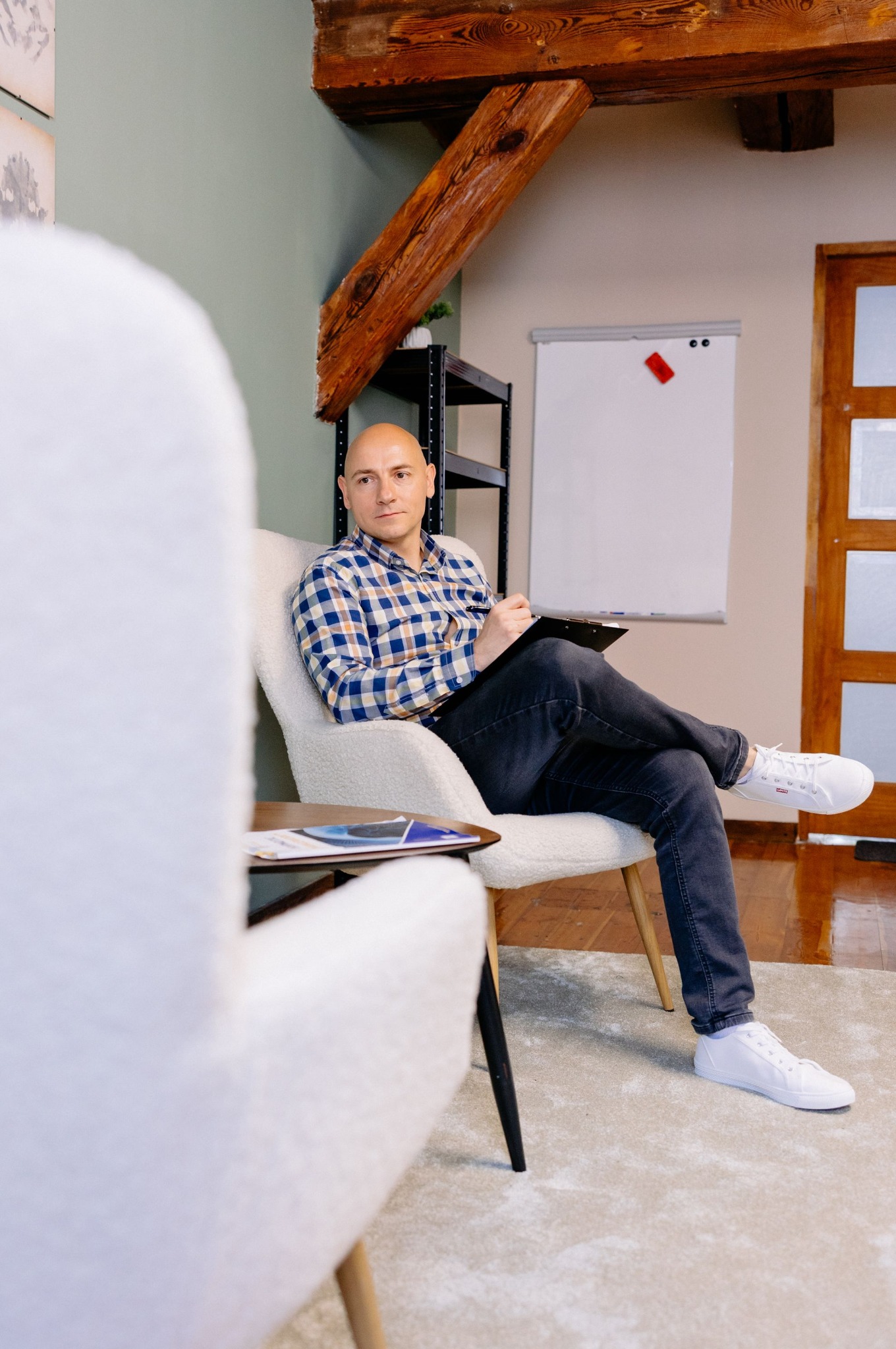 Mężczyzna w kraciastej koszuli siedzi w fotelu, trzymając notatnik na tle tablicy i drewnianych elementów wystroju wnętrza.