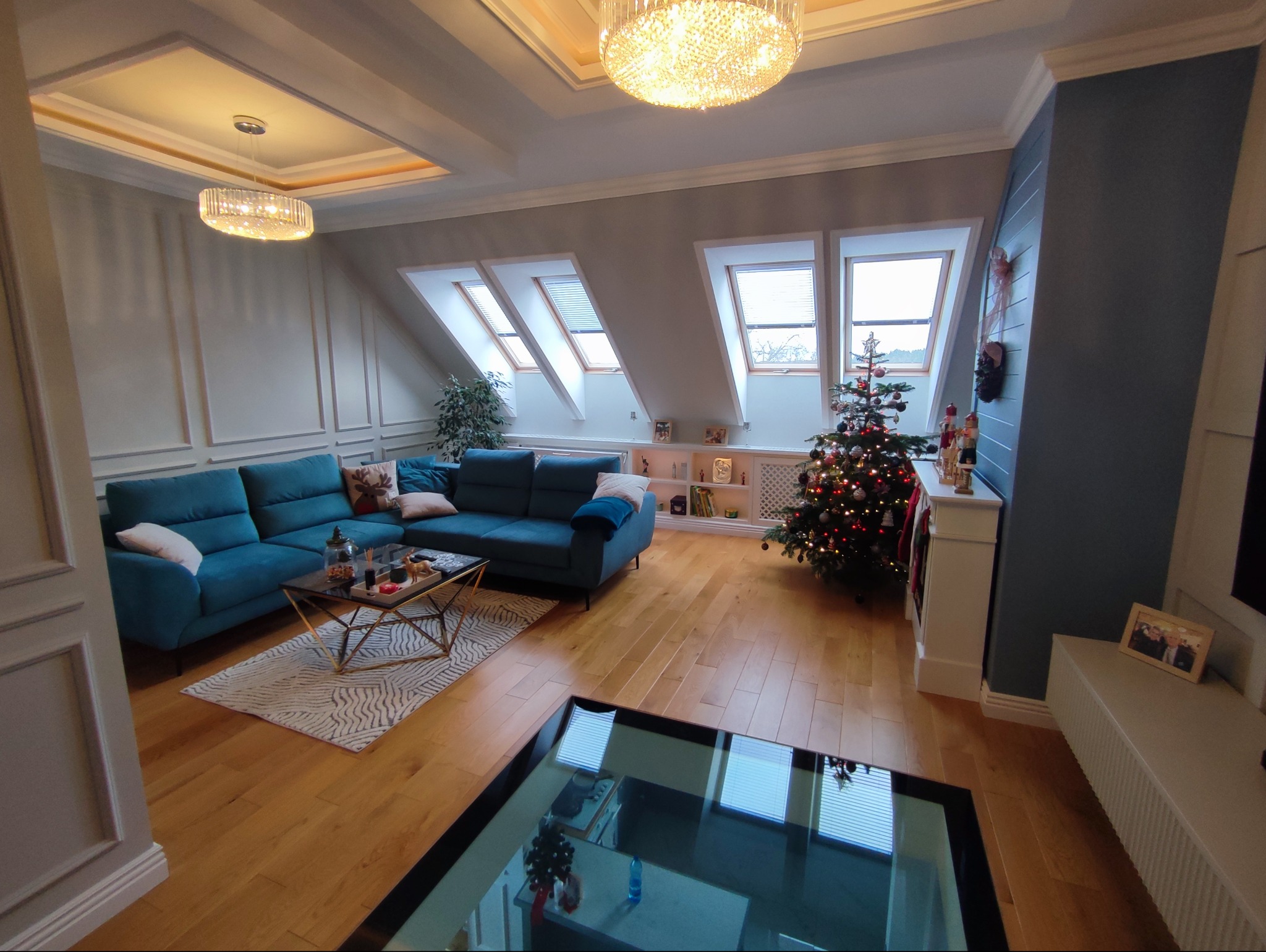 Salon na poddaszu z turkusową sofą, choinką i oknami dachowymi. Parkiet z jasnego drewna i szklana podłoga dodają wnętrzu nowoczesnego charakteru.