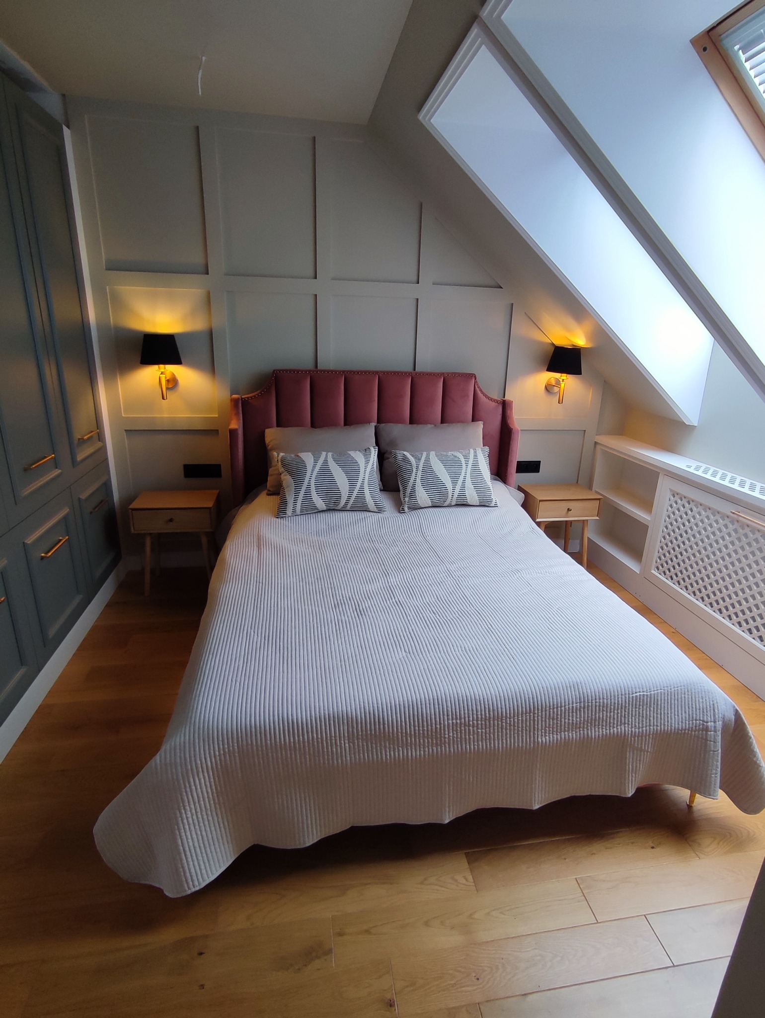 Sypialnia na poddaszu z łóżkiem z bordowym zagłówkiem, szafkami nocnymi i lampkami. Skosy z oknami dachowymi i zabudową grzejnika. Drewniana podłoga.
