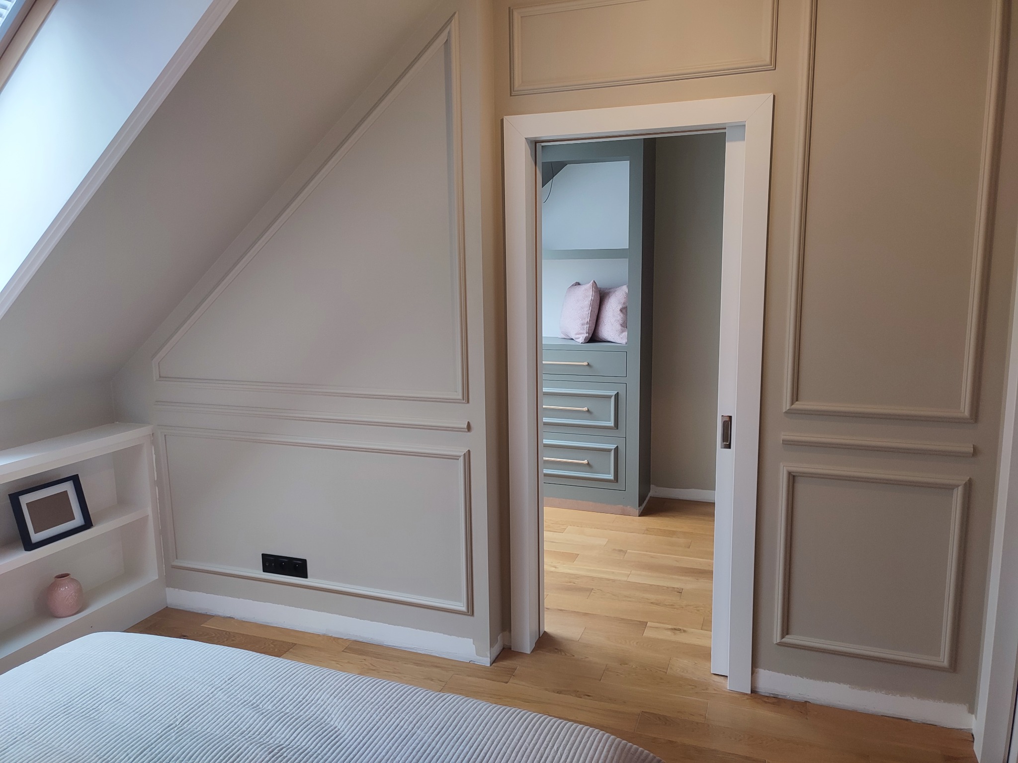 Pokój na poddaszu z drewnianą podłogą i ścianami z dekoracyjnymi listwami w odcieniach beżu. Widok na garderobę przez otwarte drzwi.