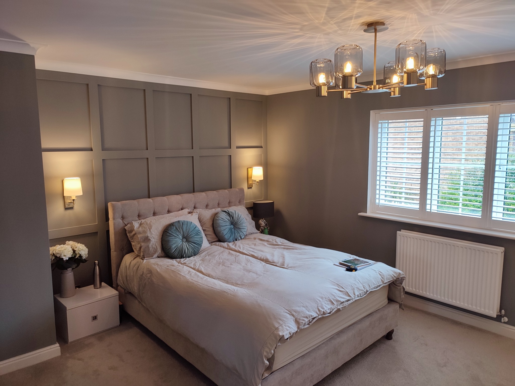 Elegancka sypialnia w odcieniach szarości z dekoracyjną sztukaterią ścienną, beżowym łóżkiem i nowoczesną lampą sufitową. Wnętrze doświetlone naturalnym światłem.