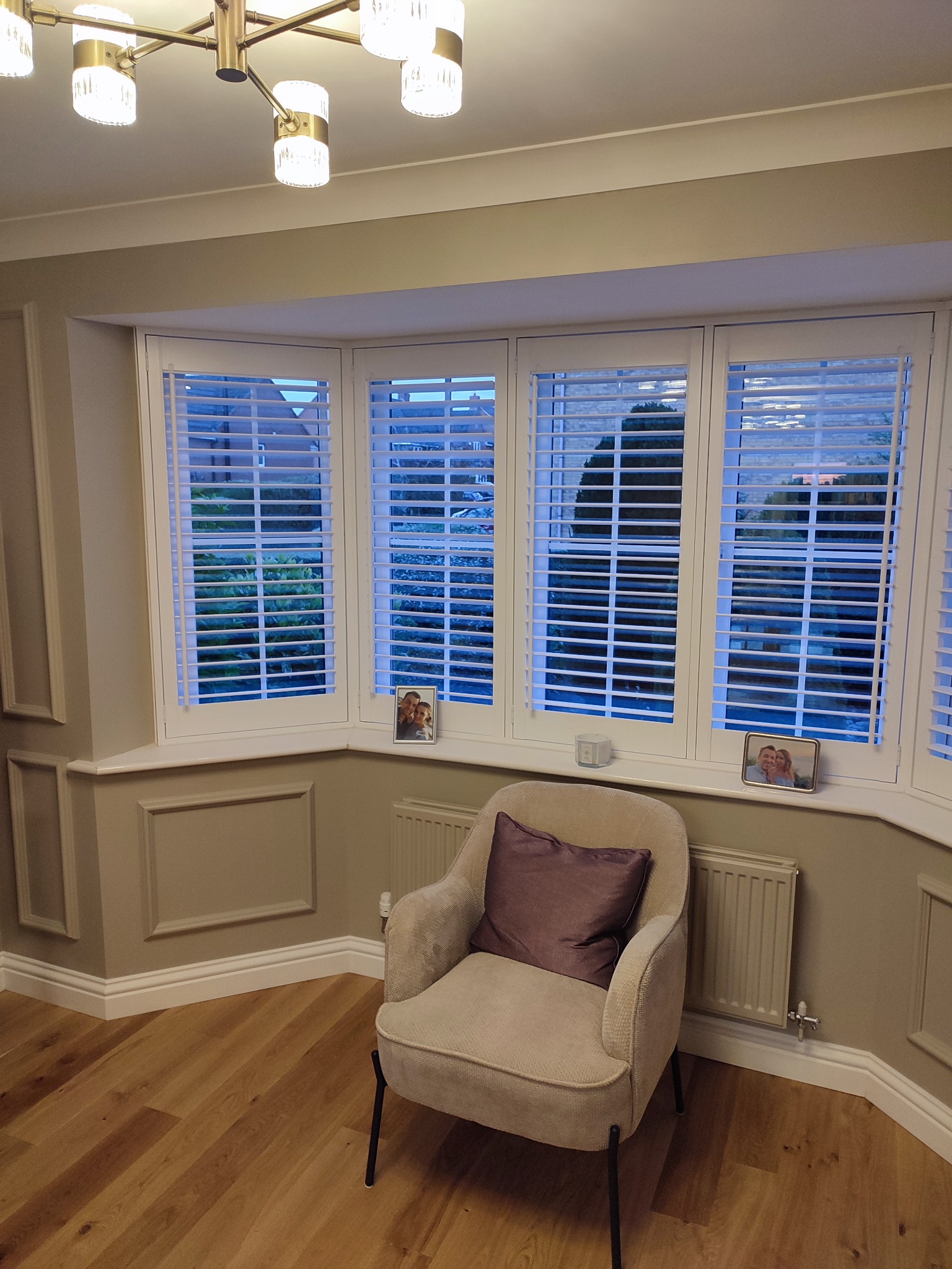 Elegancki pokój z szarymi ścianami, drewnianą podłogą i oknem z białymi żaluzjami. Beżowy fotel z fioletową poduszką stoi obok grzejnika.