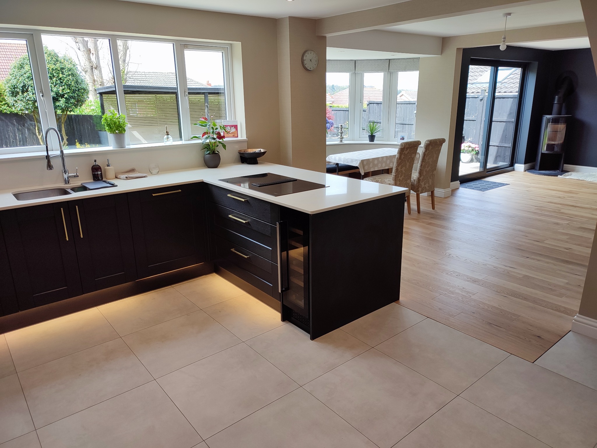 Nowoczesna kuchnia z ciemnymi szafkami, białym blatem i wyspą. Widoczne okno, stół z krzesłami i kominek. Jasna podłoga z płytek i drewniana podłoga w salonie.