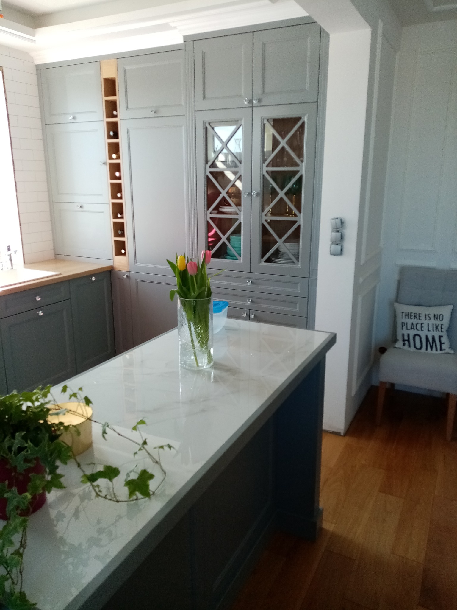 Elegancka kuchnia w szarościach: zabudowa z przeszkleniami, marmurowy blat wyspy z kwiatami, drewniana podłoga i dekoracyjna ściana z krzesłem.