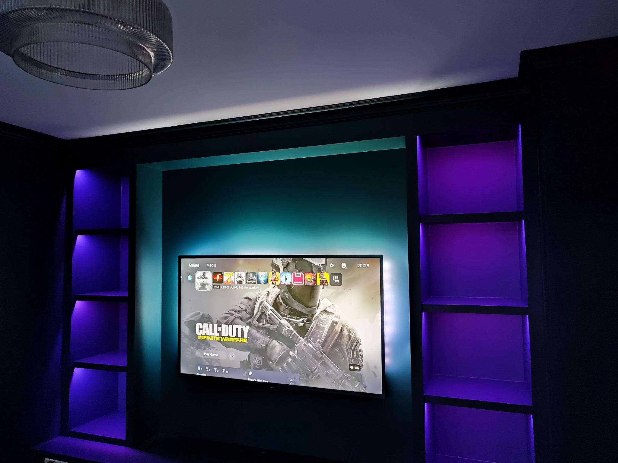 Nowoczesne wykończenie wnętrza: zabudowa TV z półkami podświetlonymi fioletowym LED, turkusowa ściana, dekoracyjny żyrandol. Widok na ekran TV z grą Call of Duty.