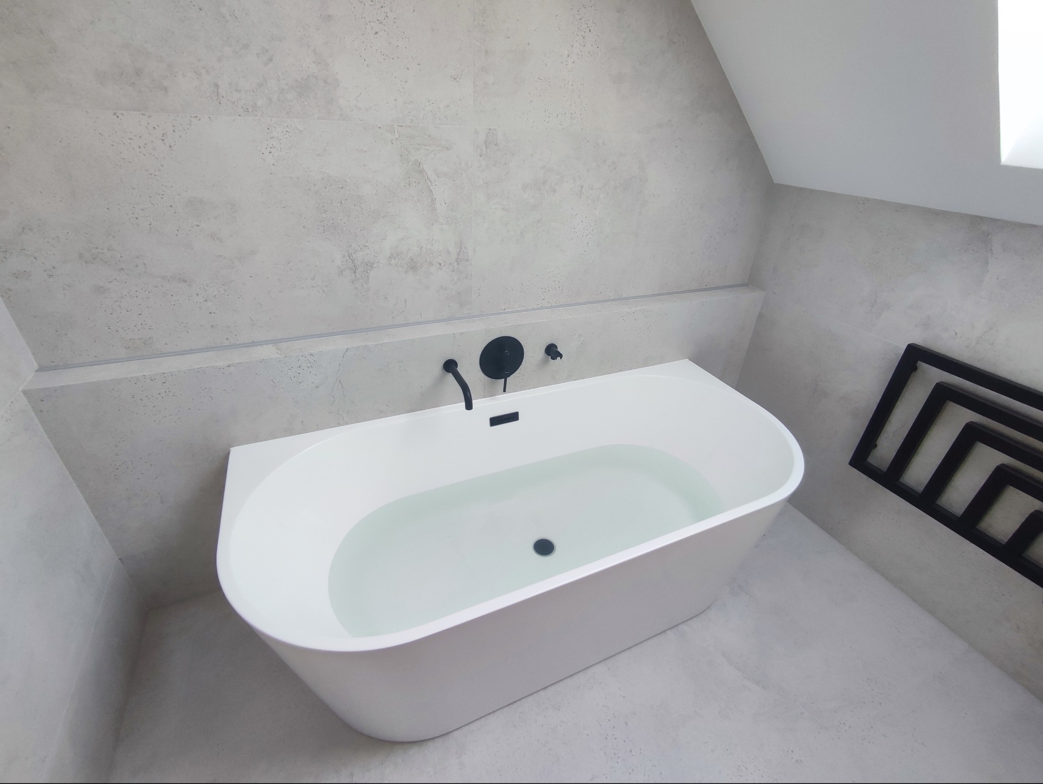 Nowoczesna łazienka na poddaszu: biała wanna wypełniona wodą, czarna armatura i grzejnik drabinkowy na tle jasnoszarych płytek imitujących beton.