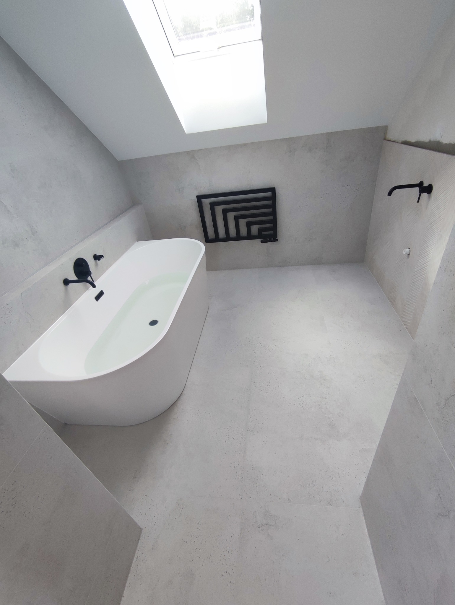 Minimalistyczna łazienka na poddaszu z białą wanną, czarną armaturą i grzejnikiem. Wnętrze w odcieniach szarości, oświetlone przez okno dachowe. Nowoczesny design.