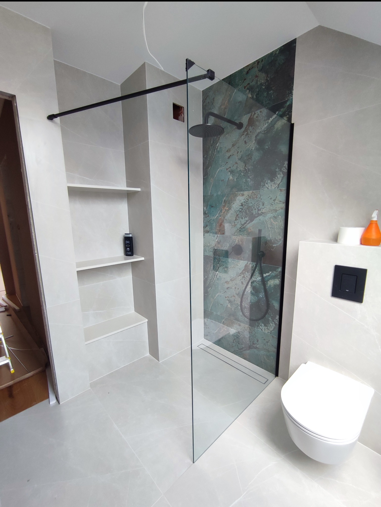 Nowoczesna łazienka z kabiną typu walk-in, wykończona płytkami w odcieniach szarości i zieleni. W tle widoczne półki wnękowe i podwieszana toaleta.