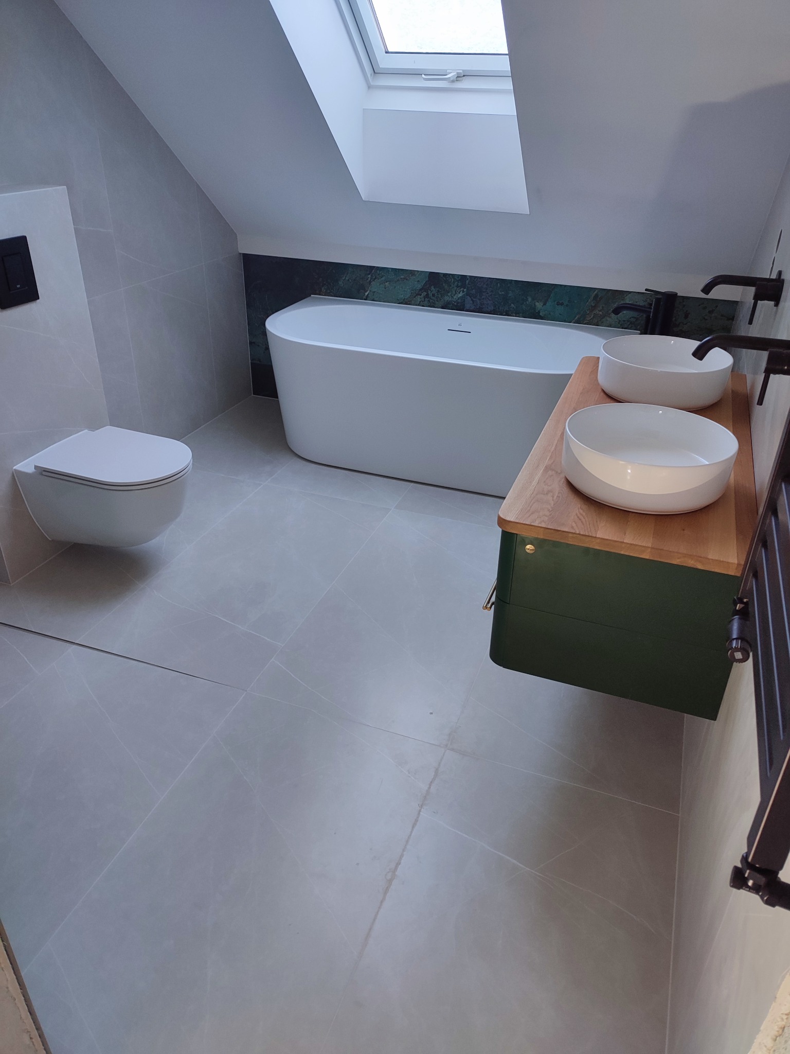 Nowoczesna łazienka na poddaszu z wolnostojącą wanną, umywalkami nablatowymi na drewnianym blacie i szarą płytką podłogową. Okno dachowe doświetla wnętrze.