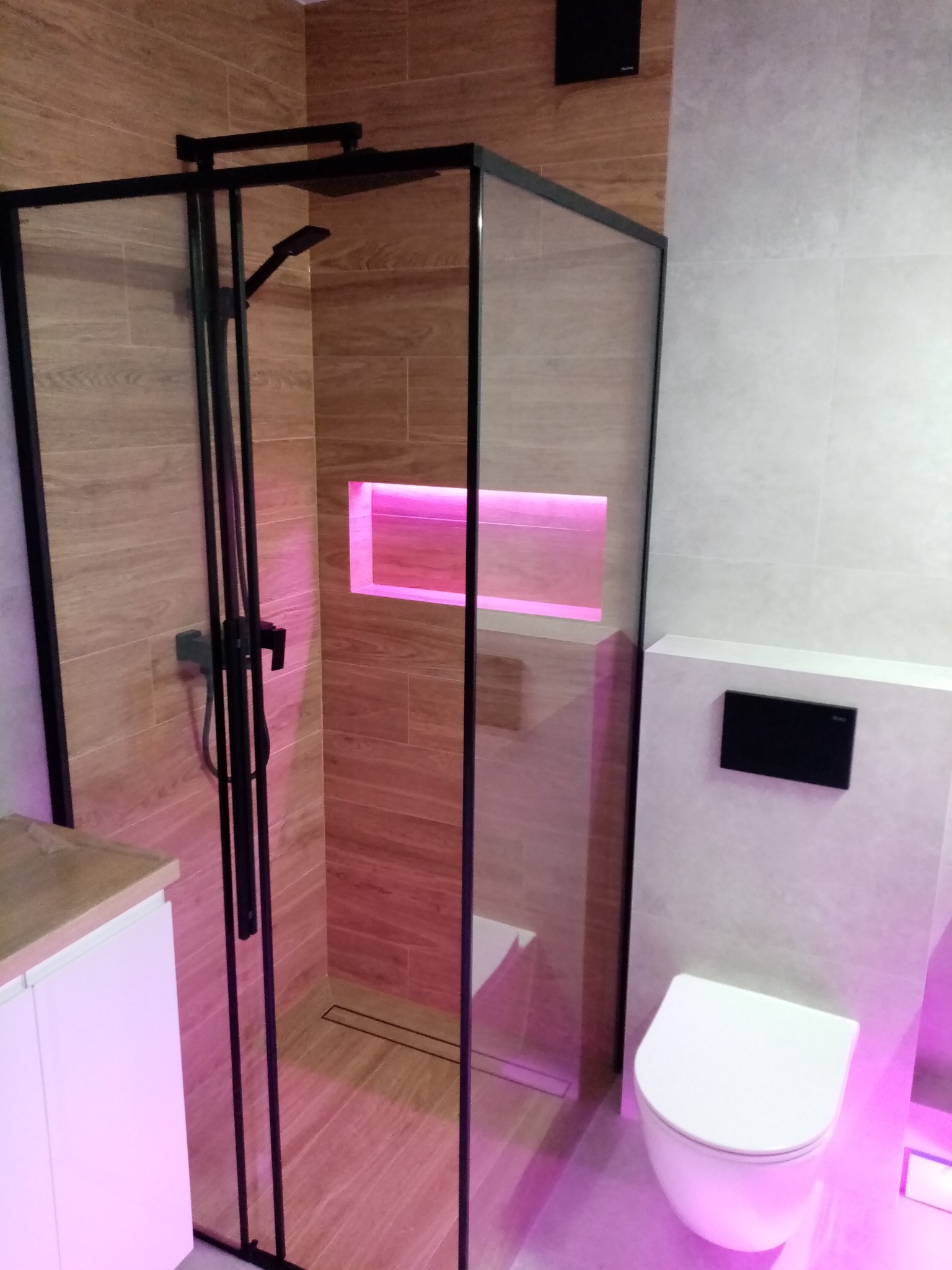 Nowoczesna łazienka z kabiną prysznicową typu walk-in, wykończona płytkami imitującymi drewno i białymi płytkami. Oświetlenie LED podkreśla detale.