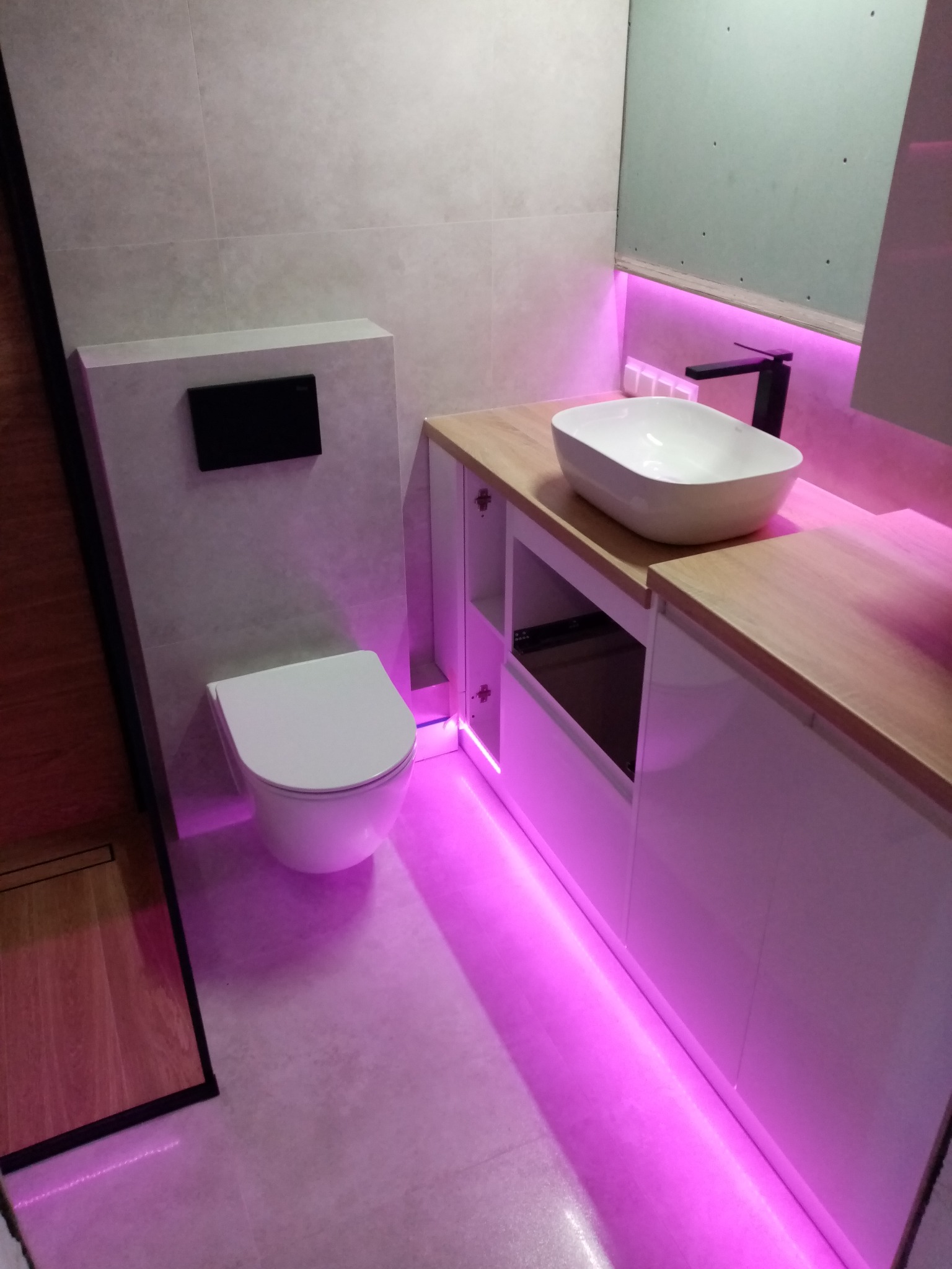 Nowoczesna łazienka z podświetleniem LED: umywalka nablatowa, wisząca toaleta i drewniany blat. Minimalistyczny design z fioletową poświatą.