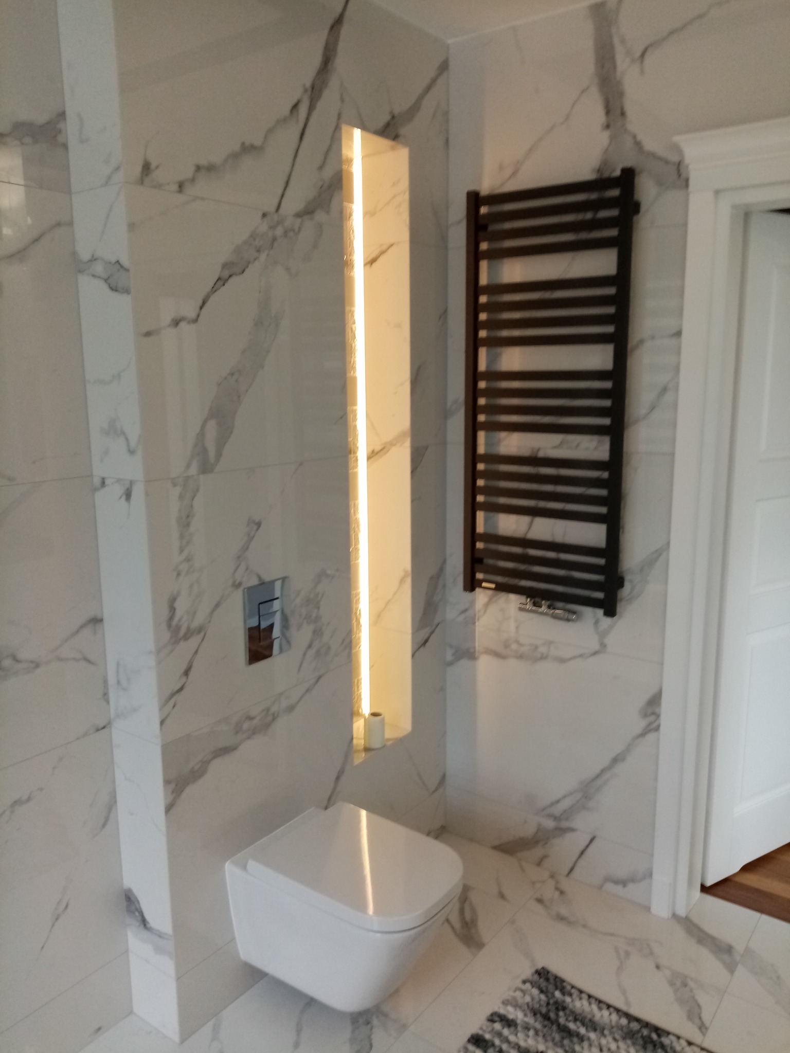 Biała łazienka z marmurowymi płytkami, podwieszaną miską WC, grzejnikiem drabinkowym i podświetlaną wnęką. Minimalistyczny design, nowoczesne wykończenie.