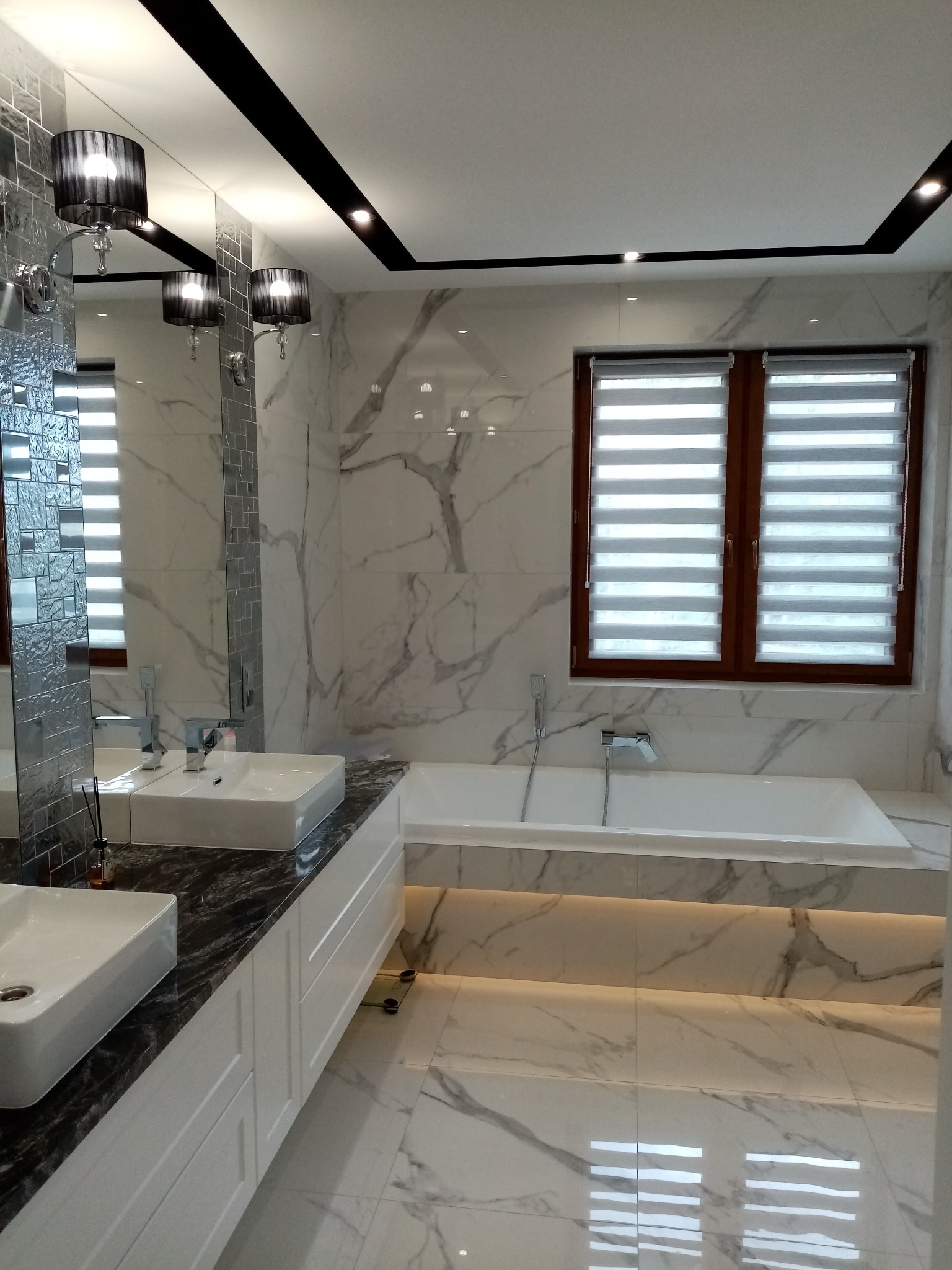 Elegancka łazienka z marmurowymi płytkami, podwójną umywalką na blacie z czarnego kamienia i podświetlaną wanną. Okno z żaluzjami w drewnianej ramie.