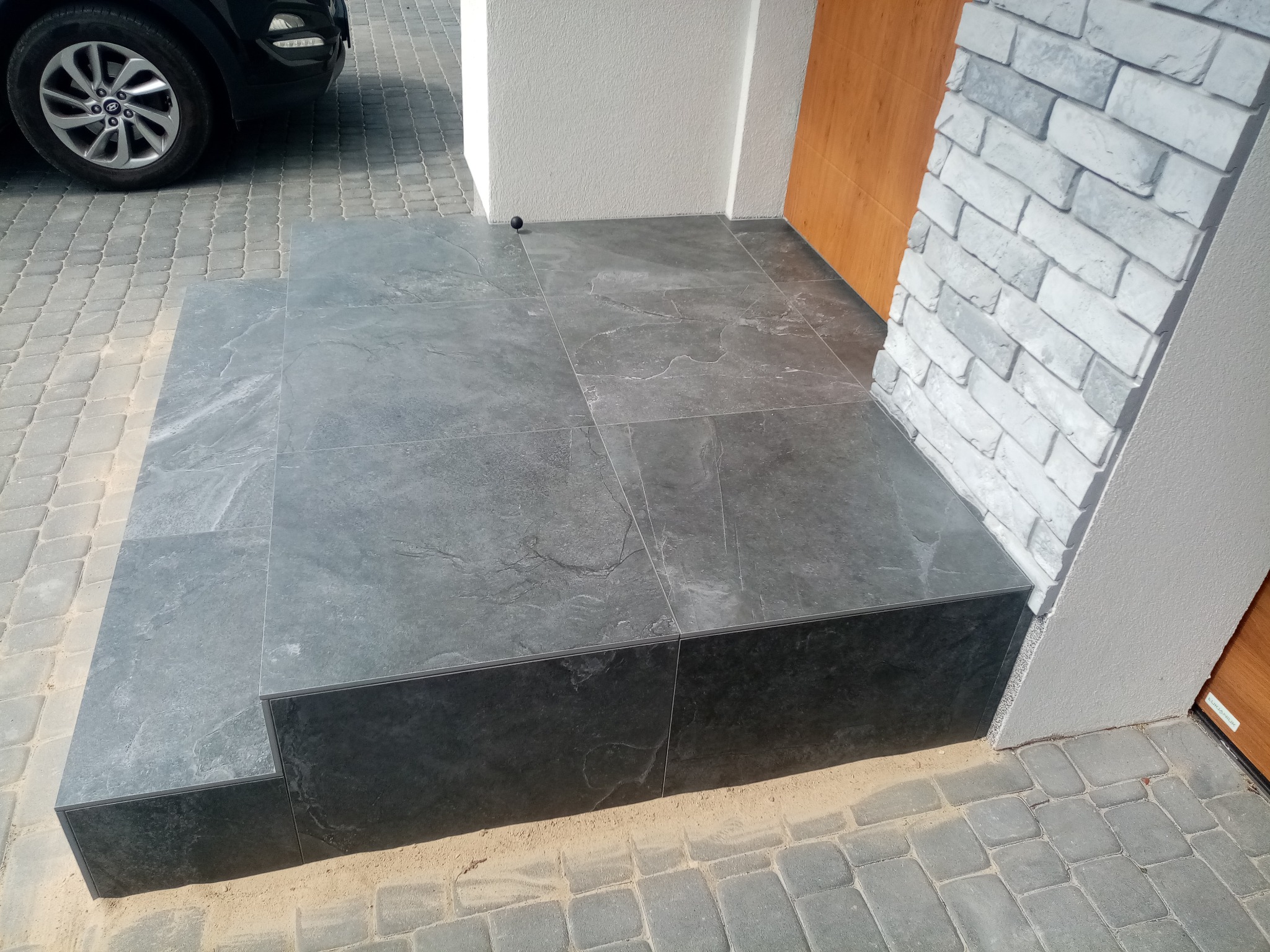 Wykończone płytkami schody wejściowe do budynku, z widoczną okładziną z cegły dekoracyjnej na filarze. Ułożona kostka brukowa przed schodami.