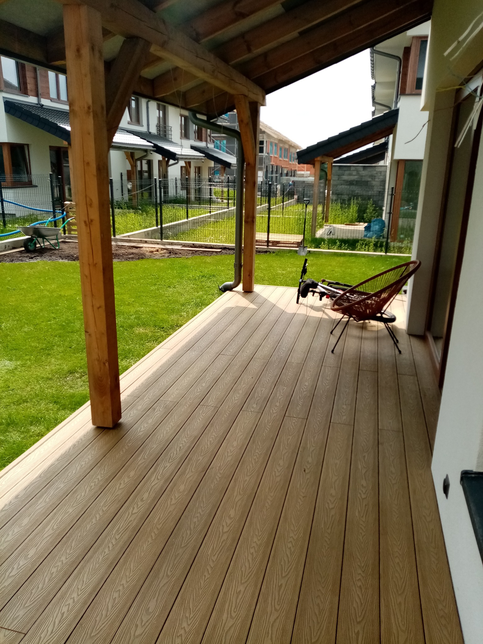 Nowoczesny taras z deski kompozytowej, zadaszony drewnianą konstrukcją. Widoczny trawnik i zabudowa szeregowa w tle. Krzesło z wikliny na tarasie.