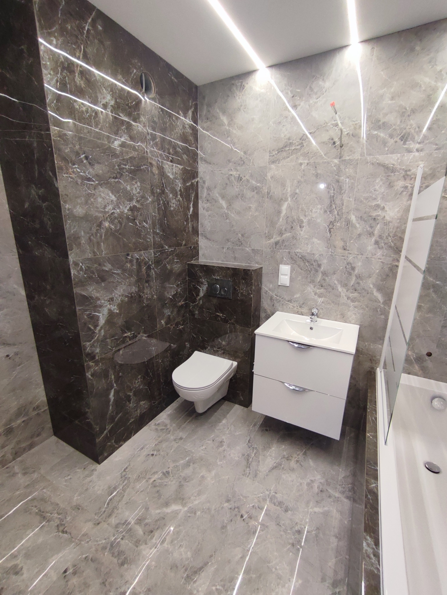 Nowoczesna łazienka z szarymi płytkami imitującymi marmur, białą szafką z umywalką i podwieszaną toaletą. Oświetlenie LED podkreśla wzór płytek.