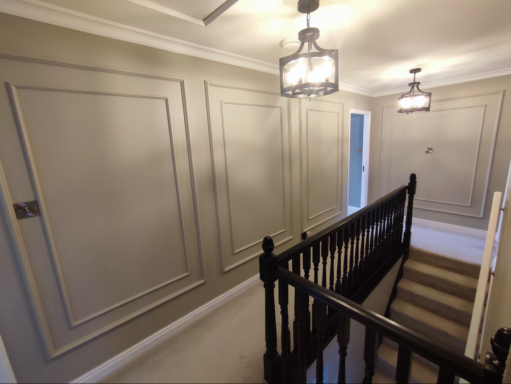 Korytarz po remoncie z panelami ściennymi w kolorze szarym, czarną balustradą schodów i nowoczesnym oświetleniem sufitowym. Widok na odnowione wnętrze.