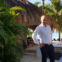 Mężczyzna w białej koszuli pozuje na tle tropikalnego krajobrazu z palmami i chatą krytą strzechą, z widokiem na błękitne morze w oddali.
