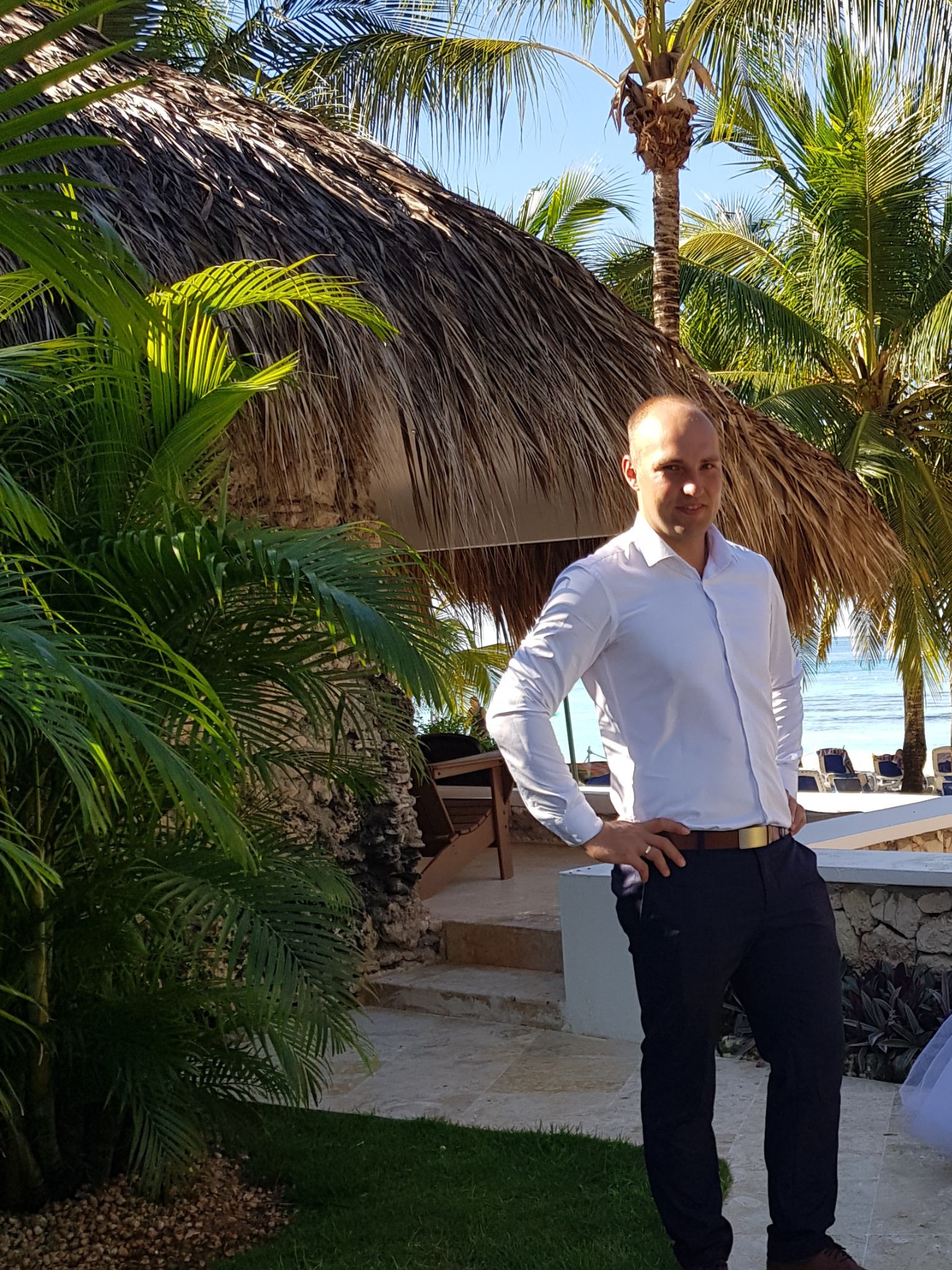 Mężczyzna w białej koszuli pozuje na tle tropikalnego krajobrazu z palmami i chatą krytą strzechą, z widokiem na błękitne morze w oddali.