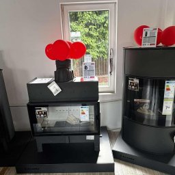 Usługi Zduńskie Dawid Oskwarek - Wnętrze salonu z trzema czarnymi kominkami marki Kratki, ozdobionymi czerwonymi balonami i ulotkami informacyjnymi, ustawionymi na tle okna z zielenią.