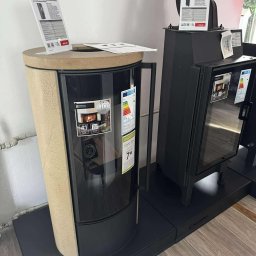 Usługi Zduńskie Dawid Oskwarek - Nowoczesne piece wolnostojące marki Kratki: okrągły, beżowy model z szybą i czarny piec z szybą, oba z etykietami energetycznymi i cenami. Ekspozycja w salonie.