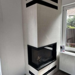 Usługi Zduńskie Dawid Oskwarek - Narożny kominek z czarnymi, poziomymi akcentami, obudowany jasnym tynkiem, z widocznym paleniskiem i szybą. Obok okno z widokiem na zieleń.