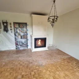 Biały, kaflowy kominek z paleniskiem w salonie w trakcie wykańczania. Widoczny parkiet i lampa w stylu retro. Usługi zduna w Kolsku.