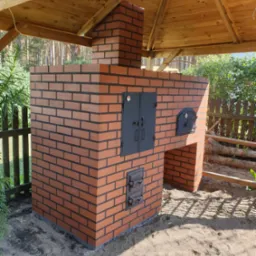 Murowany piec chlebowy z cegły klinkierowej pod drewnianą altaną, z czarnymi drzwiczkami i wędzarnią, otoczony zielenią i piaskiem.