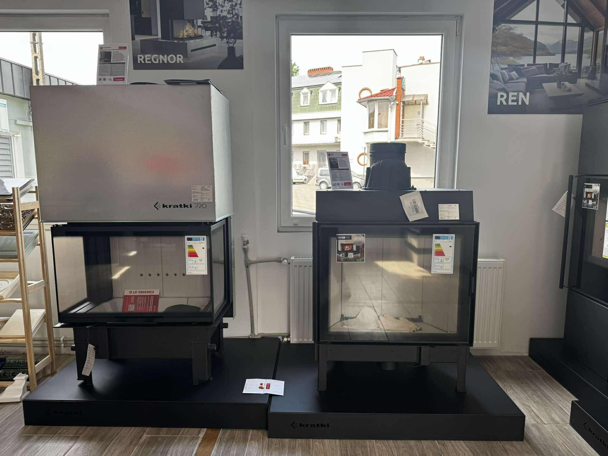 Dwa różne modele wkładów kominkowych marki Kratki, stojące obok siebie na ekspozycji w salonie. Jeden model srebrny, drugi czarny, oba z szybą.