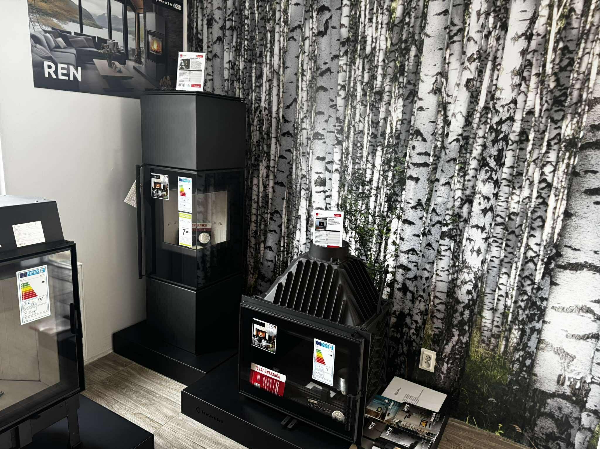 Dwa czarne kominki w salonie z tapetą w brzozy. Na kominkach naklejki z certyfikatami i danymi technicznymi. W tle plakat z kominkiem i logo Kratki.