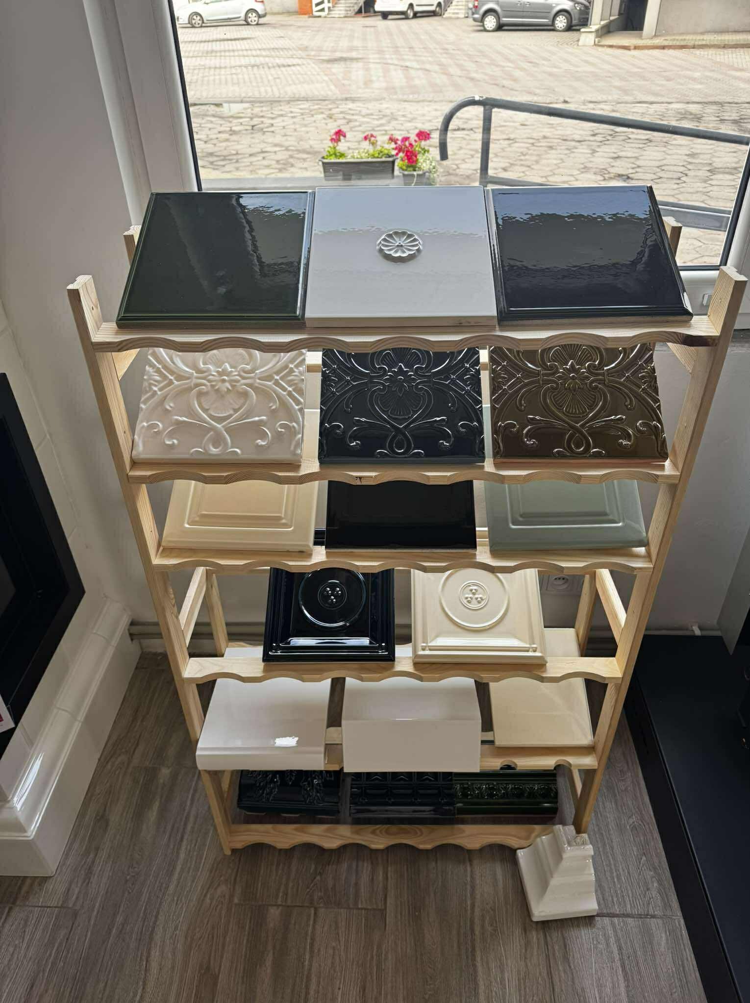 Ekspozycja kafli zdobionych, o różnych kolorach i wzorach, umieszczonych na drewnianym regale w jasnym wnętrzu. Widoczne płytki z połyskiem i reliefami.