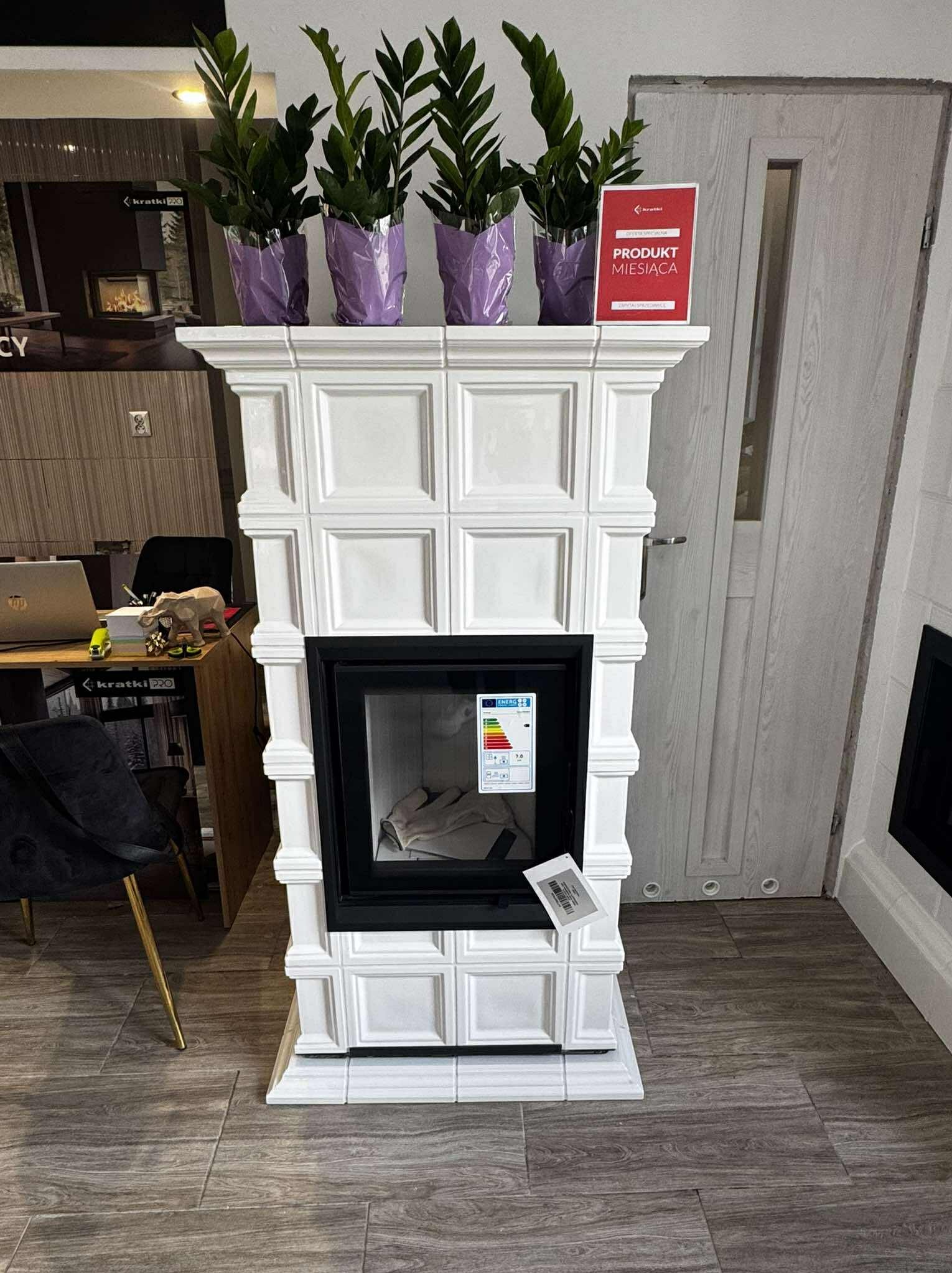 Biały piec kaflowy z czarnym wkładem, na którym stoją cztery rośliny w fioletowych osłonkach. Obok pieca widoczna tabliczka 'Produkt Miesiąca'.