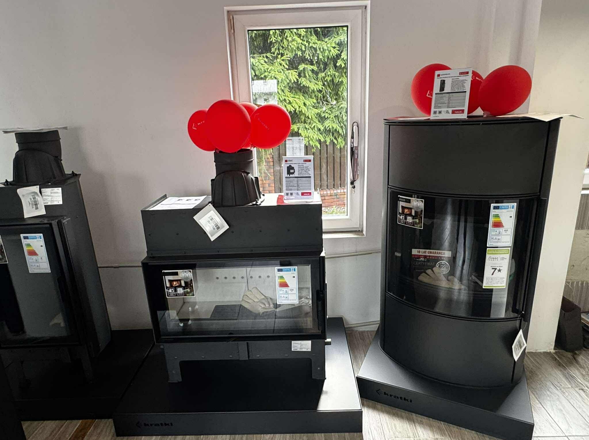 Wnętrze salonu z trzema czarnymi kominkami marki Kratki, ozdobionymi czerwonymi balonami i ulotkami informacyjnymi, ustawionymi na tle okna z zielenią.