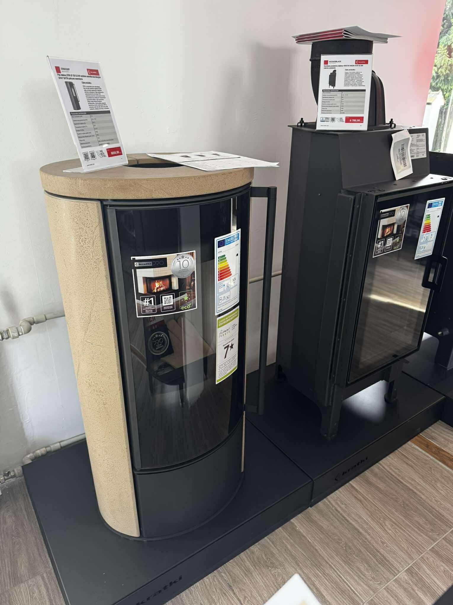 Nowoczesne piece wolnostojące marki Kratki: okrągły, beżowy model z szybą i czarny piec z szybą, oba z etykietami energetycznymi i cenami. Ekspozycja w salonie.
