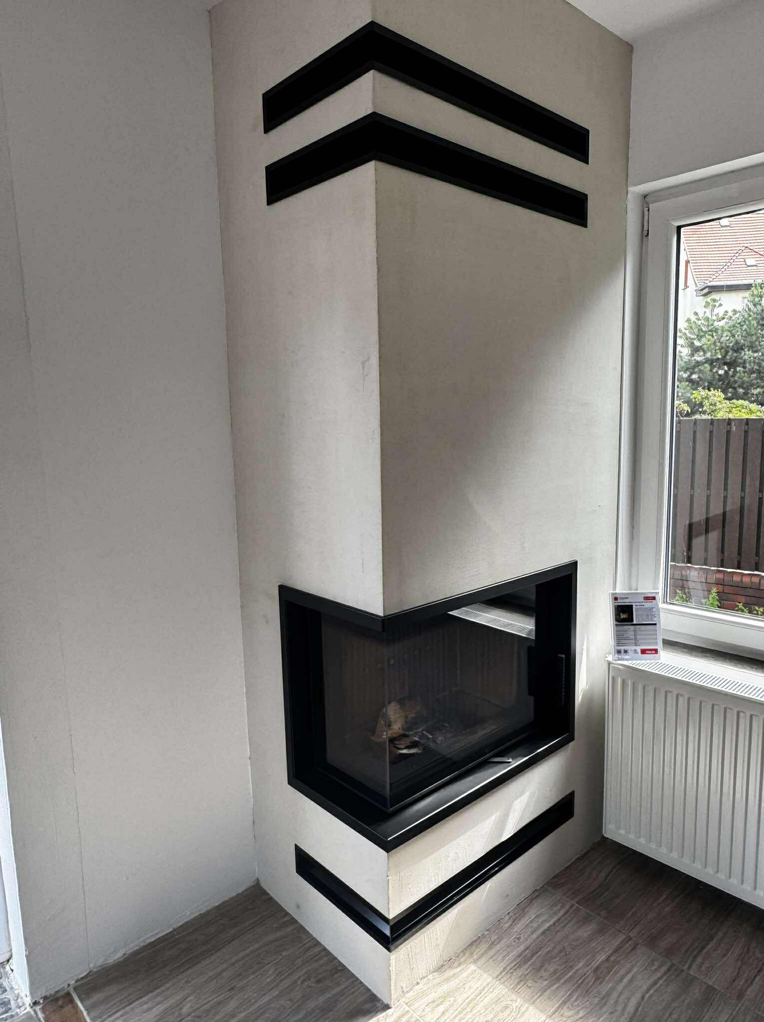 Narożny kominek z czarnymi, poziomymi akcentami, obudowany jasnym tynkiem, z widocznym paleniskiem i szybą. Obok okno z widokiem na zieleń.
