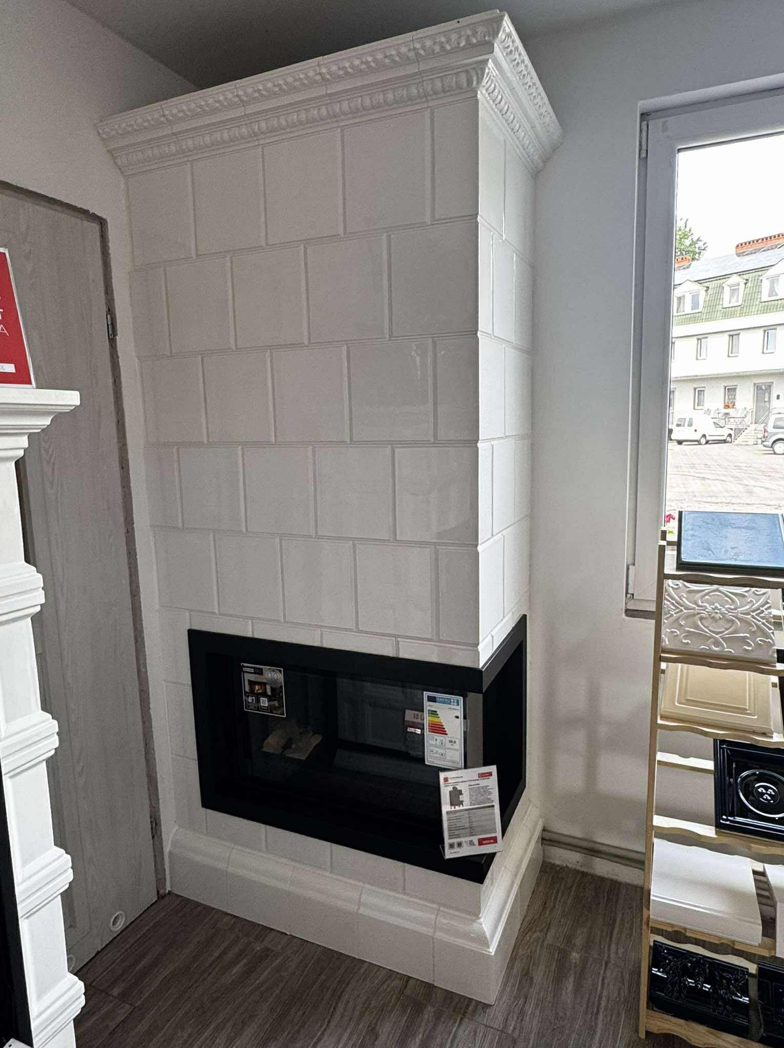 Biały, kaflowy piec z czarnym wkładem kominkowym, ozdobiony gzymsem. Ustawiony w rogu pomieszczenia, obok okna i regału z elementami dekoracyjnymi.