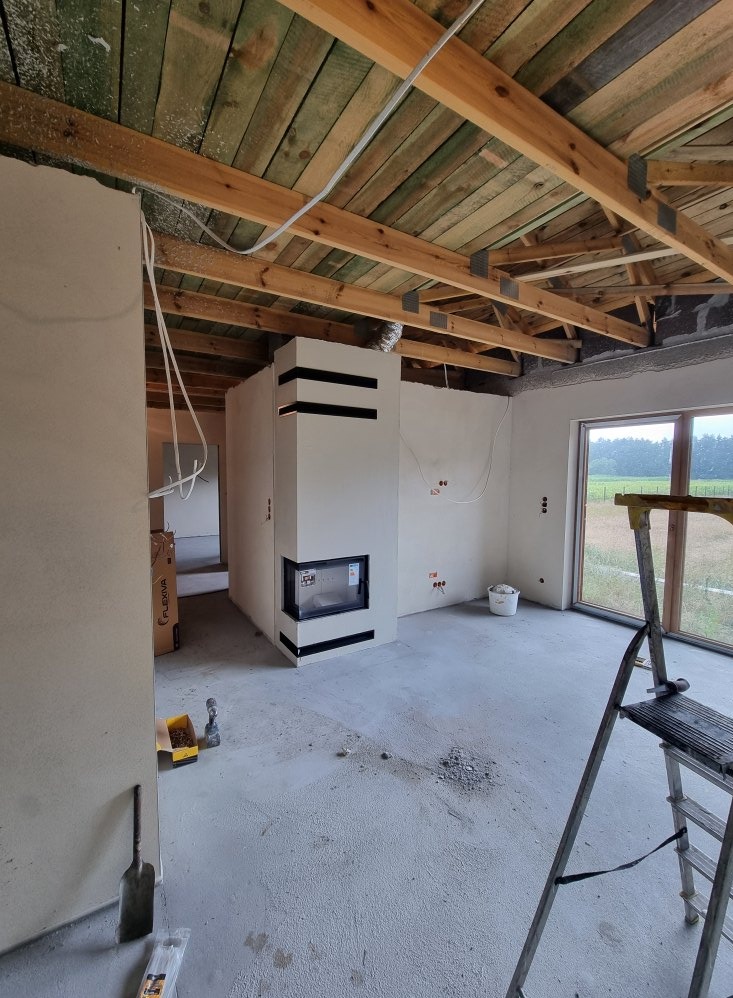 Nowoczesny, biały kominek z czarnymi akcentami w surowym wnętrzu domu w trakcie budowy. Widoczne drewniane elementy konstrukcyjne dachu i drabina.