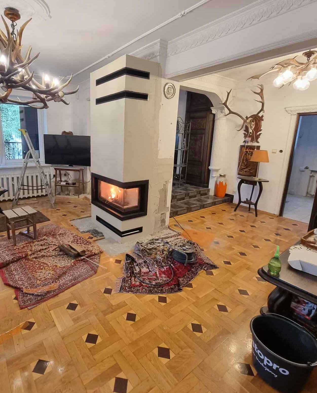 Wnętrze z kominkiem o geometrycznej obudowie w trakcie prac wykończeniowych, widoczne narzędzia i zabezpieczone podłogi. Dekoracje z poroża jelenia.